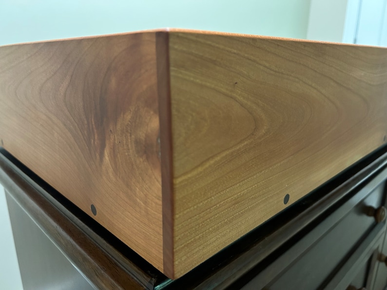 May include: Close-up of a wooden box with a natural wood finish. The box has a smooth surface and is made of solid wood. The box is sitting on a dark brown surface.
