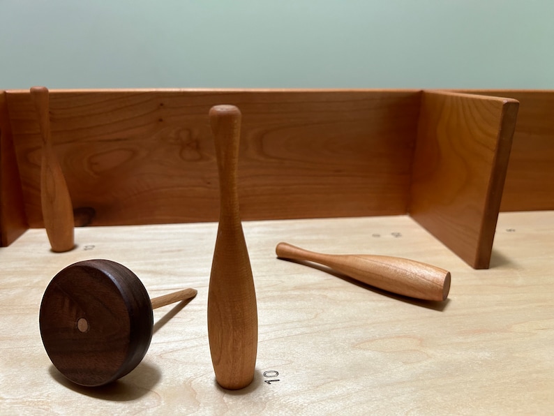 May include: A wooden game set with a brown wooden wheel and several wooden bowling pins. The pins are arranged on a light wood surface with numbers marked on the surface.