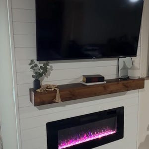 May include: A contemporary living room arrangement with a large black television mounted above a wooden shelf. Beneath, a fireplace with a black frame shows a vibrant pink and purple flame effect. The wall is white with horizontal wood panelling.