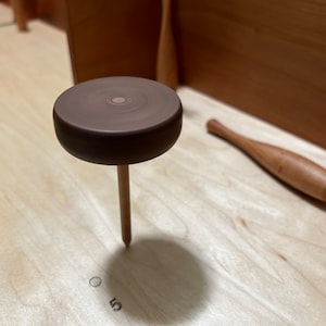 May include: A wooden game piece with a brown top and a wooden peg. The piece is sitting on a light wood surface.