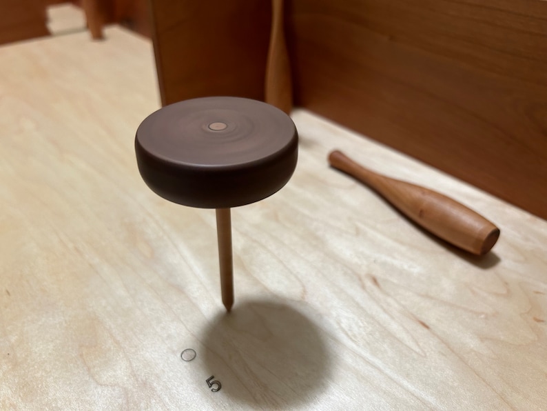 May include: A wooden game piece with a brown top and a wooden peg. The piece is sitting on a light wood surface.