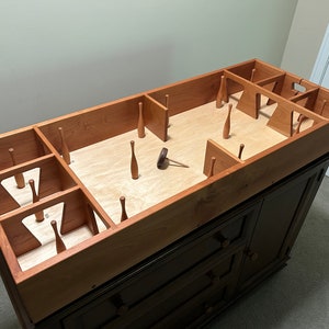 May include: A wooden game with a rectangular playing surface and multiple wooden pins. The game is set up on a dark brown dresser with three drawers. The game is made of light brown wood and has a natural wood finish.