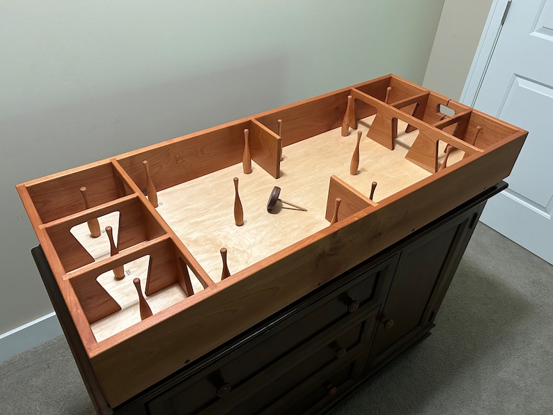 May include: A wooden game with a rectangular playing surface and multiple wooden pins. The game is set up on a dark brown dresser with three drawers. The game is made of light brown wood and has a natural wood finish.