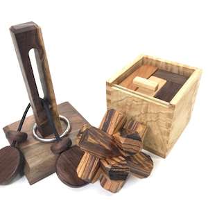 May include: Wooden brain teaser puzzles in various shapes and designs. Includes a dark walnut puzzle with a ring and string, a wooden box puzzle, and a cluster of geometric wooden pieces. The puzzles are on a white background.