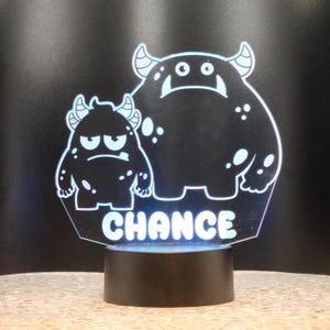 May include: A white acrylic night light with two black cartoon monsters and the name "CHANCE" printed below. The light is on a black base.