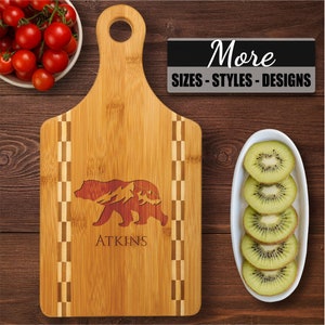 May include: A personalized bamboo cutting board with a checkered border. The board features a laser-engraved design of a bear with mountains in the background. The text "ATKINS" is engraved below the bear.