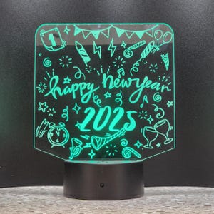 May include: A green acrylic LED night light with the words "happy new year 2025" and festive illustrations. The light sits on a black base and is designed for holiday decor.