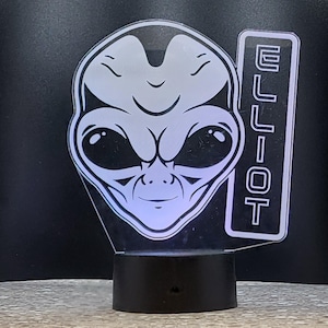 May include: A white alien face with large eyes and a small mouth, with the name "ELLIOT" written vertically on the side. The alien face is a 3D printed night light with a black base.