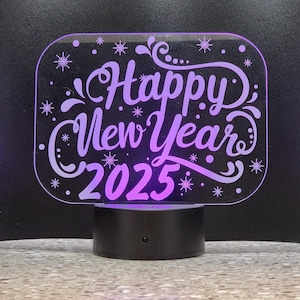 May include: A purple illuminated sign with the text "Happy New Year 2025" in a decorative font. The sign is on a black base.
