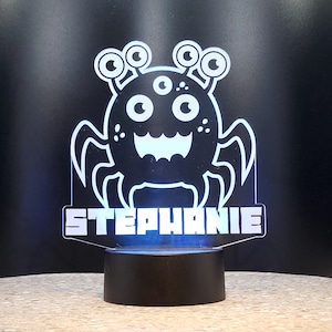 May include: A white LED night light shaped like a cartoon monster with five eyes and eight legs. The name "STEPHANIE" is printed below the monster.
