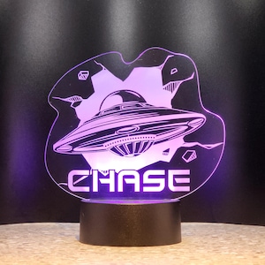 May include: A purple-lit UFO-themed night light with the name "CHASE" illuminated in white. The design features a detailed illustration of a flying saucer against a backdrop of stylized mountains and geometric shapes. The base is black.