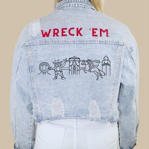 May include: A light blue denim jacket with distressed detailing. The back of the jacket features the text "WRECK 'EM" in red lettering and a black and white embroidered design of a cowboy, a building, and a horse.