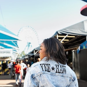 May include: Light blue denim jacket with the text "YEE HAW" in black block letters and a star graphic. The jacket is acid-washed and has a classic design. The image is taken outdoors, with a Ferris wheel in the background.
