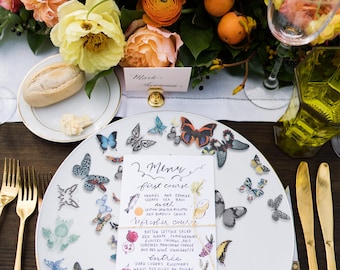 May include: A beautifully set table with a white plate decorated with butterfly decals. A menu card is on the plate, and gold cutlery is arranged to the side. The table is adorned with a floral centerpiece of peach and yellow roses.