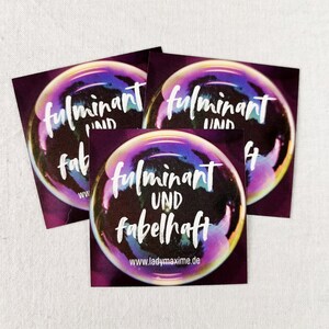 Könnte beinhalten: Drei quadratische Aufkleber mit lila Hintergrund und einem Blasendesign. Die Blase hat schillernde Farben und die Worte "fulminant und fabelhaft" in weißer Schrift. Die Website-Adresse www.ladymaxime.de ist ebenfalls sichtbar.