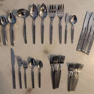 May include: A collection of stainless steel flatware featuring a geometric pattern on the handles. The set includes various spoons, forks, knives, a ladle, and a serving spoon, all arranged in rows on a light-colored surface.