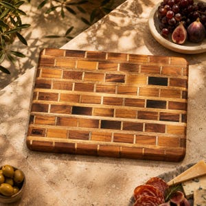 May include: A rectangular wooden cutting board featuring a brick-like pattern made of alternating light and dark brown wood segments. The board has a smooth, flat surface and a small rectangular logo stamp in the bottom left corner of the wood.