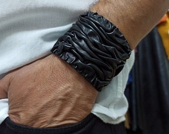 May include: A black leather bracelet with a wrinkled, textured design. The bracelet is worn on a person's wrist.