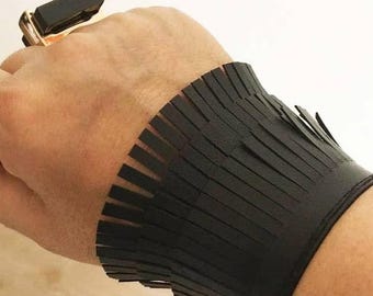 Könnte beinhalten: Schwarzes Lederarmband mit Fransen-Design. Das Armband ist breit und umschließt das Handgelenk. Ein goldener Ring mit einem großen schwarzen rechteckigen Stein ist ebenfalls zu sehen.
