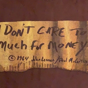 Puede incluir: Una camiseta marr&oacute;n con una imagen de papel desgarrado y amarillento de una nota manuscrita que dice "I Don't Care Too Much For Money... &copy; 1964 John Lennon/Paul McCartney".
