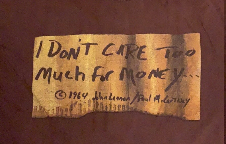 Puede incluir: Una camiseta marr&oacute;n con una imagen de papel desgarrado y amarillento de una nota manuscrita que dice "I Don't Care Too Much For Money... &copy; 1964 John Lennon/Paul McCartney".