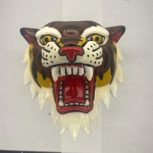 Puede incluir: Una escultura de cabeza de tigre de cerámica con fondo blanco, rayas marrones y amarillas, y una boca roja abierta con dientes blancos.