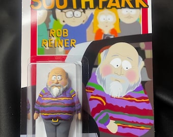 May include: A South Park Rob Reiner action figure in a clear plastic package. The figure is wearing a purple, red, and green striped sweater, gray pants, and a white beard. The packaging has the South Park logo and the text "OUT OF STOCK".