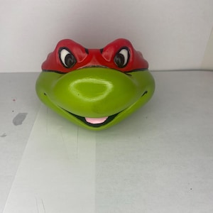 Pode incluir: Uma cabeça de personagem de Teenage Mutant Ninja Turtles de cerâmica verde e vermelha com olhos pretos e uma língua rosa.