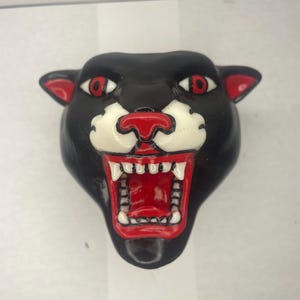 Puede incluir: Una cabeza de pantera de cerámica negra con ojos rojos y boca blanca, la pantera está gruñendo con los dientes visibles.