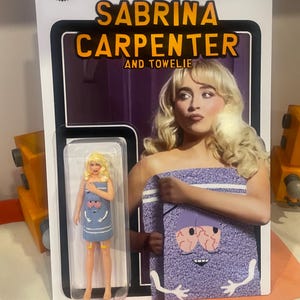 May include: A Sabrina Carpenter and Towelie action figure in a clear plastic case. The packaging features a photo of Sabrina Carpenter and the text "SABRINA CARPENTER AND TOWELIE". The figure is wearing a blue dress with a towel design.