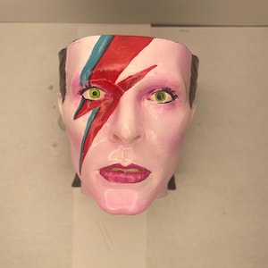 May include: A white ceramic bust of a person with a red and blue lightning bolt painted on the face. The person has green eyes and pink lips.