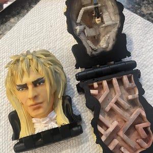 Könnte beinhalten: Ein Vintage-Spielzeug zum Thema Labyrinth mit einem geformten Gesicht und blonden Haaren. Das Spielzeug lässt sich öffnen und zeigt ein Miniatur-Labyrinth und Figuren. Das Spielzeug ist aus Kunststoff und hat ein schwarzes Scharnier.