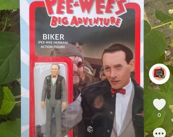 May include: A vintage-style action figure of Pee-wee Herman dressed as a biker, with a gray suit, black leather vest, and a red bow tie. The figure is packaged in a red and white box with the words "Pee-wee's Big Adventure" and "Biker (Pee-wee Herman) Action Figure".