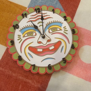 May include: A round wall clock featuring a stylized face with a white base, green and red scalloped border, and black numerals from 1 to 12. The face has blue, yellow, and red accents around the eyes and mouth, with two black clock hands centered.