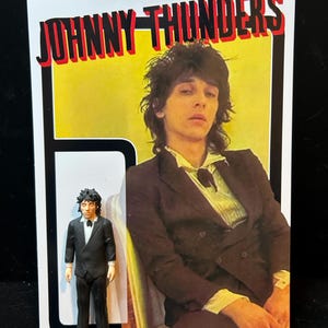 Puede incluir: Una figura de acción de Johnny Thunders con traje negro, camisa blanca y corbata negra, presentada frente a una tarjeta rectangular con una fotografía de Johnny Thunders, el texto "JOHNNY THUNDERS" y una advertencia de "CHOKING HAZARD".