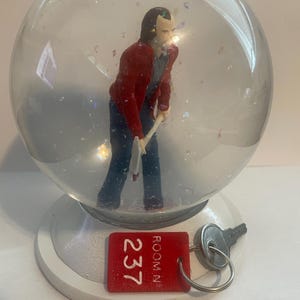 May include: A snow globe featuring a miniature figure of a man in a red jacket and blue pants holding an axe. The base is white, and a red tag reads "237" with a key attached.
