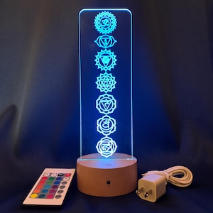 May include: A wooden base with a clear acrylic panel displaying seven chakra symbols in a vertical line. The panel is illuminated with a blue light. The base has a remote control and a power adapter.