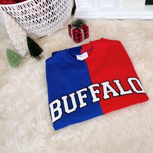 May include: A red and blue sweatshirt with the word "BUFFALO" in white letters on the front. The sweatshirt is folded and lying on a white fluffy surface.