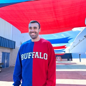 May include: A person wearing a blue and red long-sleeved shirt with the word "BUFFALO" printed in white on the front. The person is standing in front of a building with a red and blue striped awning.