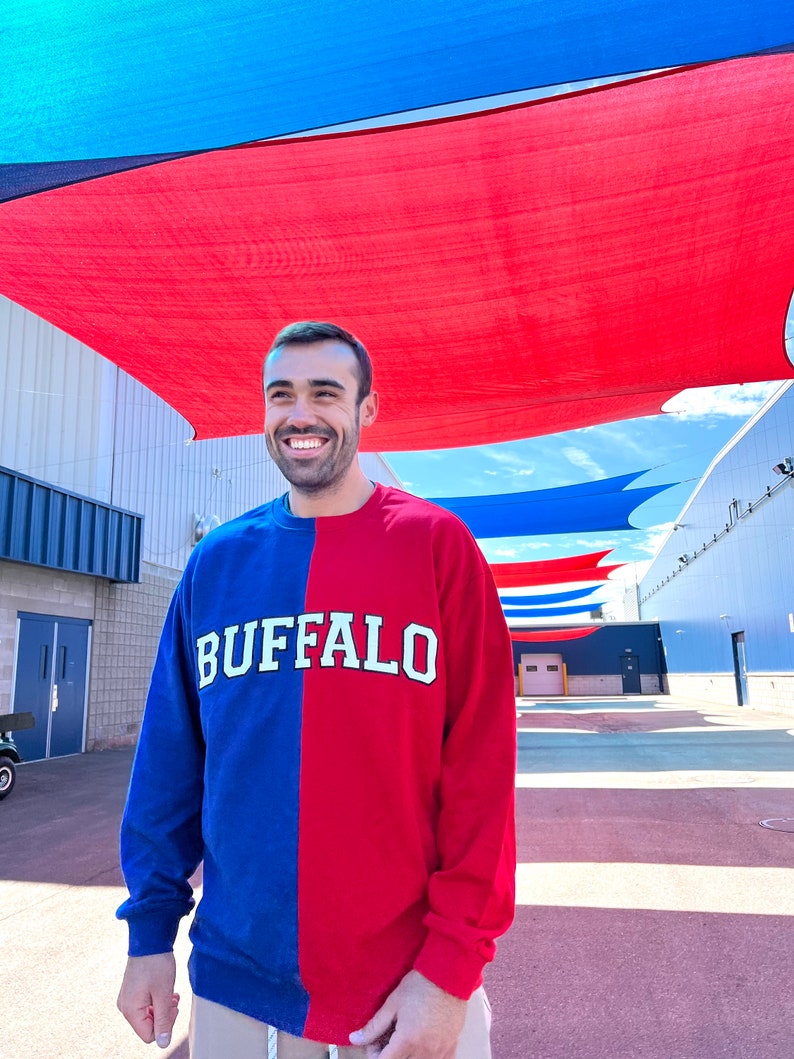 May include: A person wearing a blue and red long-sleeved shirt with the word "BUFFALO" printed in white on the front. The person is standing in front of a building with a red and blue striped awning.
