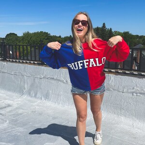 May include: A woman wearing a blue and red sweatshirt with the word "BUFFALO" printed on it. She is wearing denim shorts and white sneakers. She is standing on a rooftop with a blue sky in the background.
