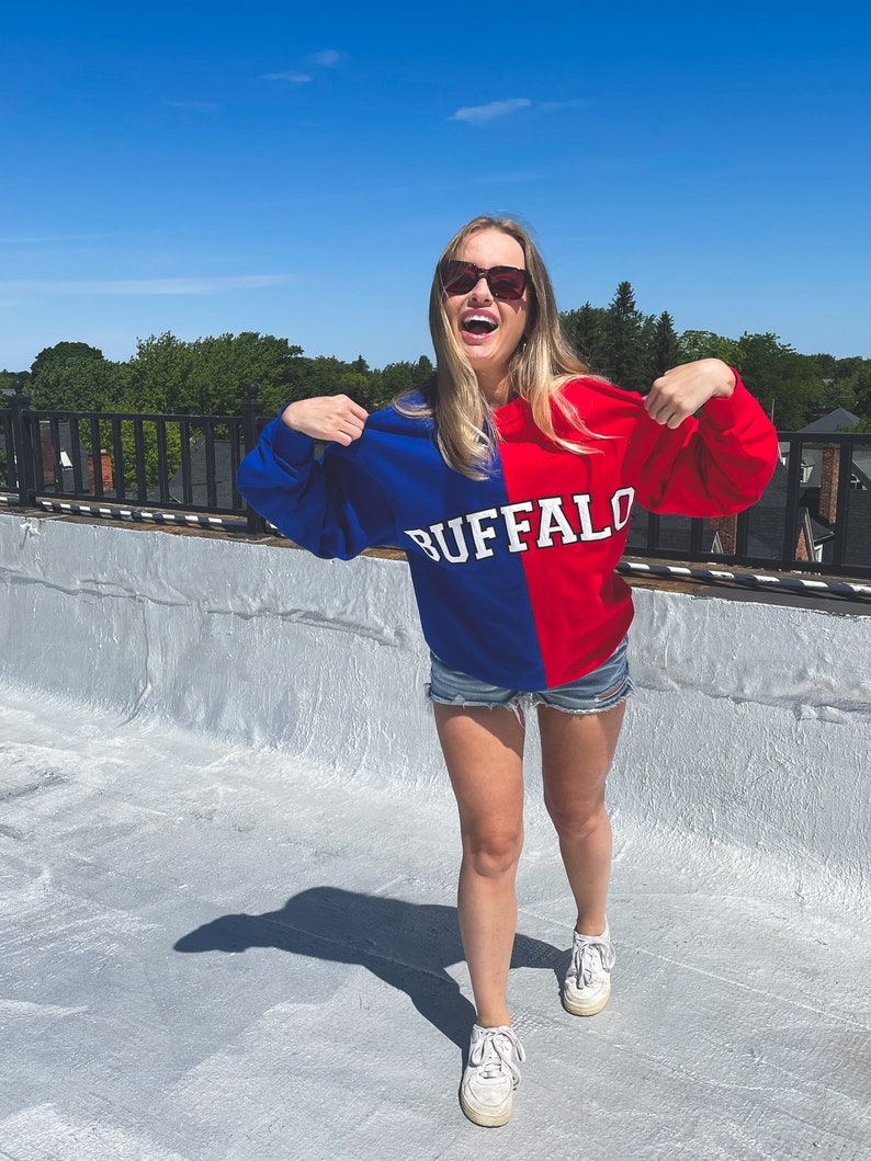 May include: A woman wearing a blue and red sweatshirt with the word "BUFFALO" printed on it. She is wearing denim shorts and white sneakers. She is standing on a rooftop with a blue sky in the background.
