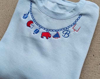 May include: A light blue crewneck sweatshirt featuring an embroidered design of a chain necklace with charms. The charms include a blue foam finger, a red football helmet, a blue football, a red buffalo, a red megaphone, a blue letter B, and a goal post.