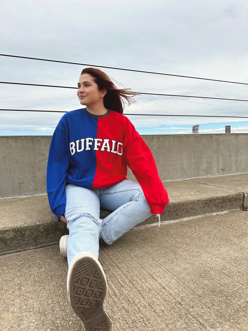 May include: A person wearing a blue and red sweatshirt with the word "BUFFALO" in white lettering. The person is sitting on a concrete surface with their legs crossed and wearing light blue jeans and white sneakers.