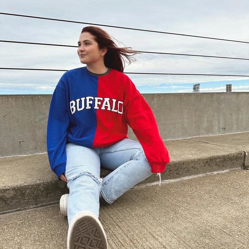 May include: A woman wearing a red and blue sweatshirt with the word "BUFFALO" printed on the front. She is sitting on a concrete surface with her legs crossed.