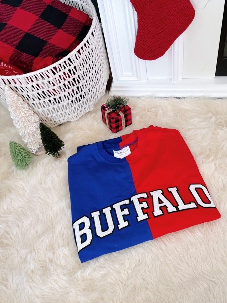 May include: A red and blue sweatshirt with the word "BUFFALO" in white letters on the front. The sweatshirt is folded and lying on a white fluffy surface.