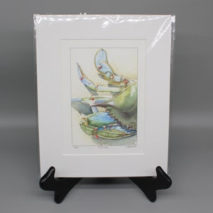 May include: A watercolor print of a blue crab, displayed in a white mat and clear plastic sleeve. The crab is depicted in shades of blue and green, with a sandy background. The print is titled "Blue Crab".