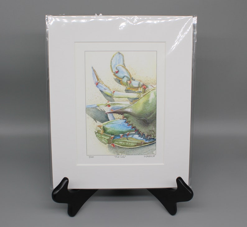 May include: A watercolor print of a blue crab, displayed in a white mat and clear plastic sleeve. The crab is depicted in shades of blue and green, with a sandy background. The print is titled "Blue Crab".