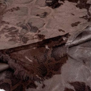May include: A dark brown animal hide featuring a textured snakeskin pattern embossed onto the surface. The material has an irregular, organic shape with raw, uneven edges and a soft, slightly fuzzy appearance across the dark brown leather.