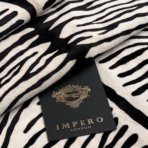 May include: A piece of fabric featuring a black and white zebra stripe pattern. A black square tag is placed on top of the fabric, displaying the gold text "IMPERO LONDON" and a gold ornate emblem above the brand name, centered on the tag.